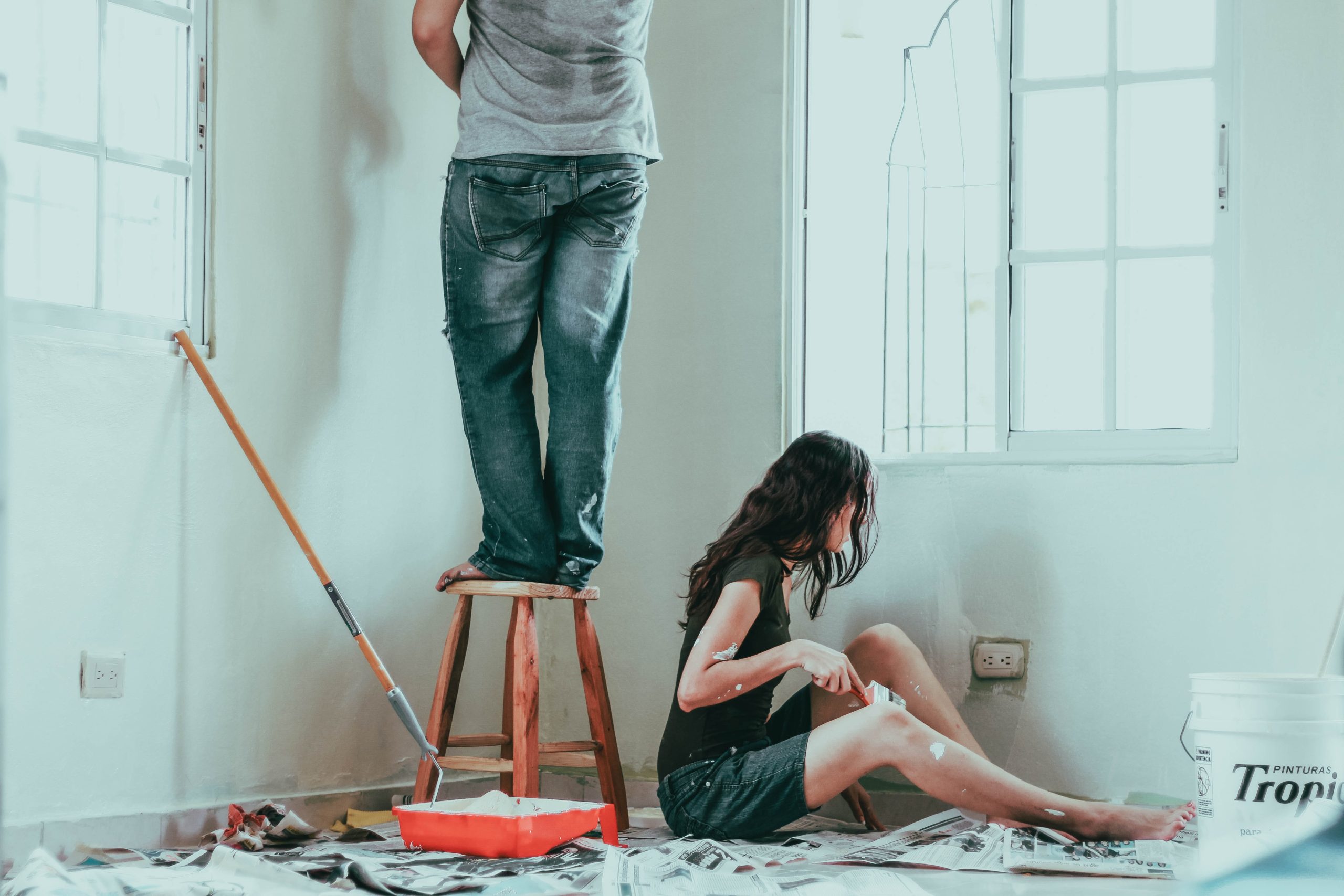 two people painting, the woman sitting on the floor while the man is standing on a stool